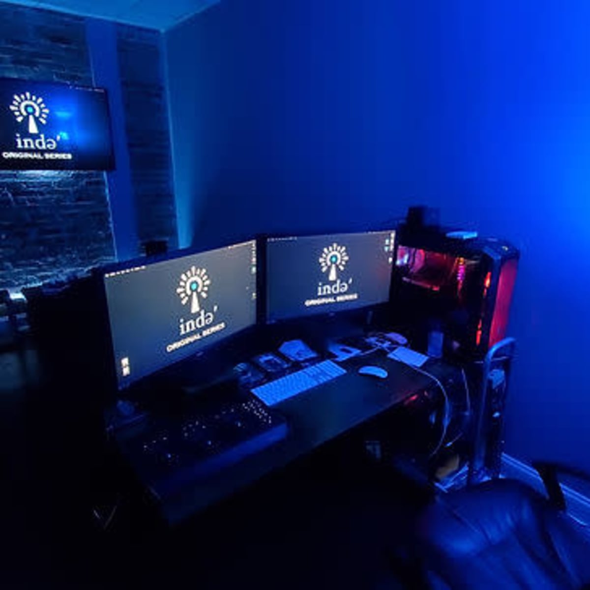 A dimly lit workspace featuring two computer monitors, a modern gaming PC, and ambient blue lighting.