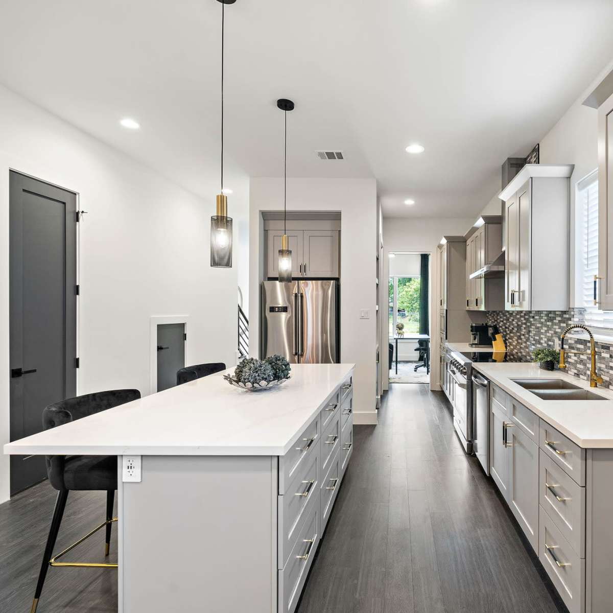 A modern kitchen features a central island with a light-colored countertop, gray cabinetry, and a mosaic tile backsplash, leading to a doorway that opens to an outdoor view.