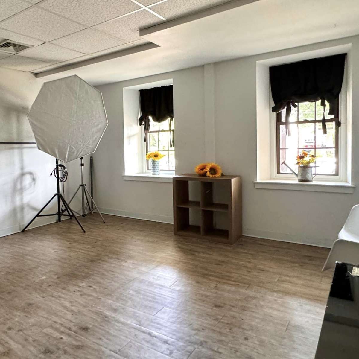 A vacant room with two windows, a light-colored floor, a photo backdrop setup, a small shelving unit, and a chair.