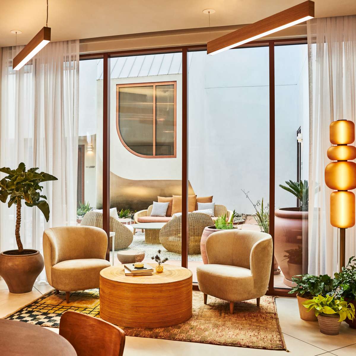 A modern living space with large windows, featuring two armchairs, a round coffee table, a plant, and a decorative lamp, all overlooking a courtyard with seating and greenery.