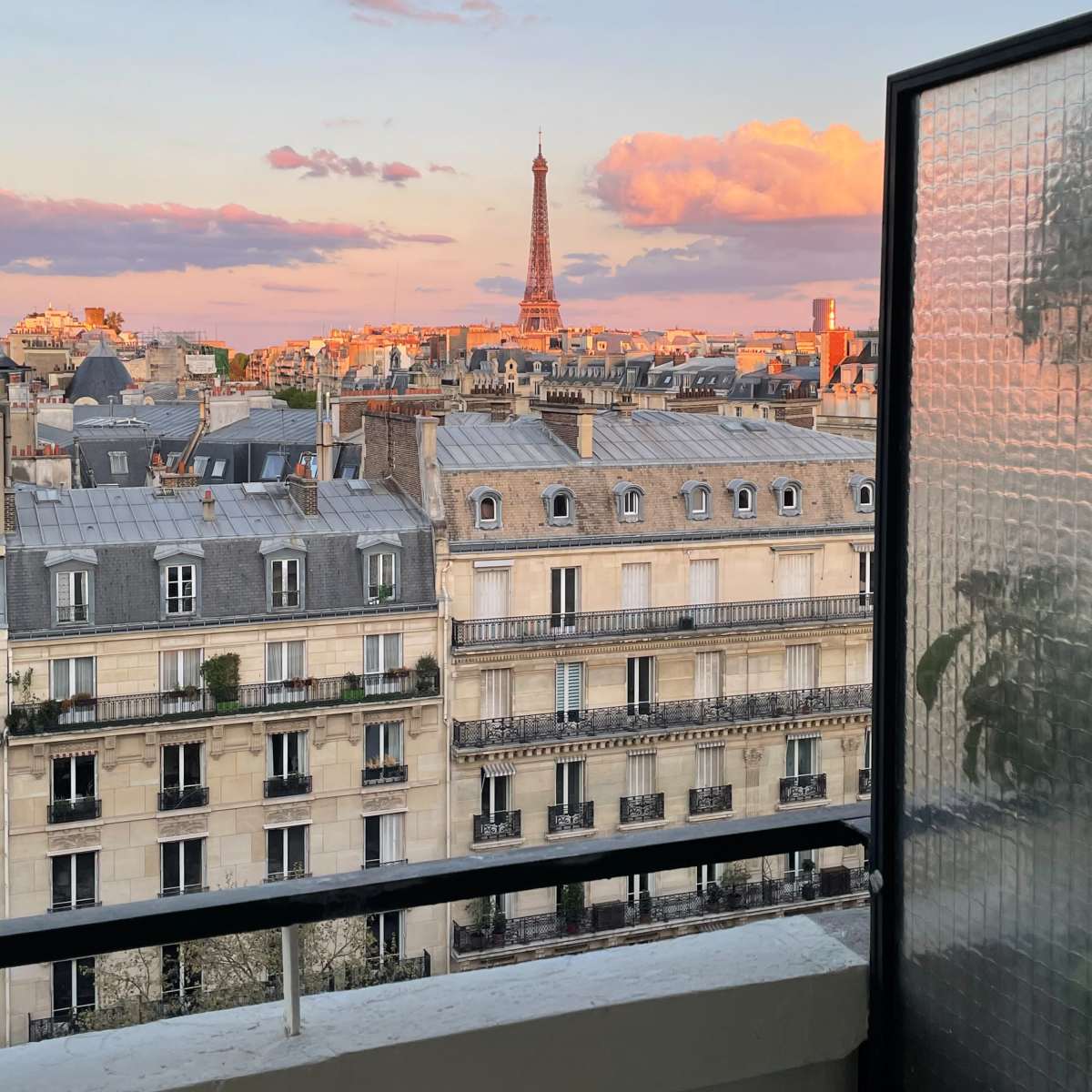 A view from a balcony overlooking Paris, with the Eiffel Tower visible in the distance against a colorful sunset sky.
