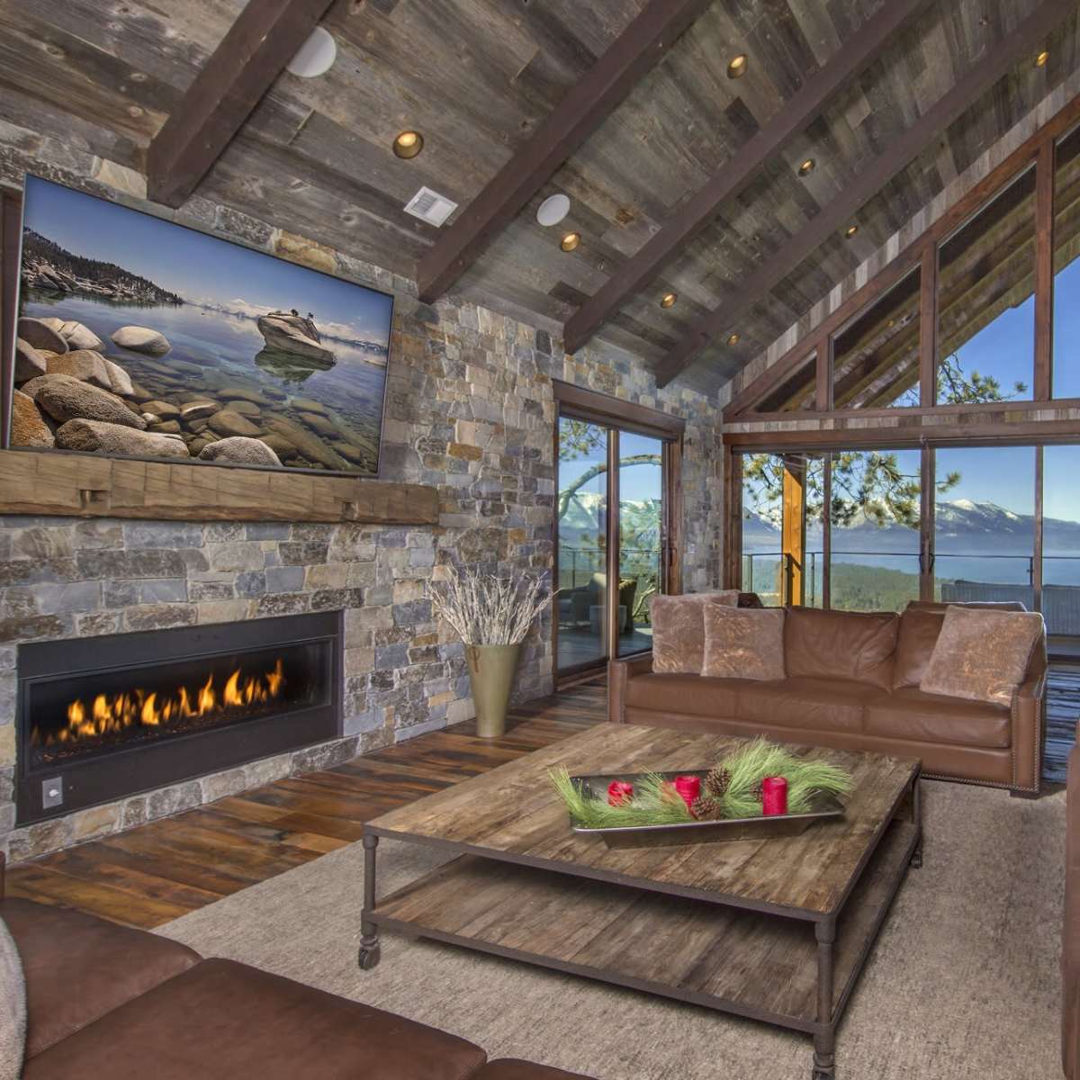 The living room features a stone fireplace, large windows with a view of the mountains, and leather furniture arranged around a coffee table.