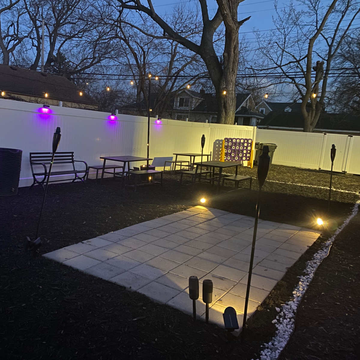 An outdoor patio area with purple lights, seating arrangements, and a paved square surrounded by tiki torches.