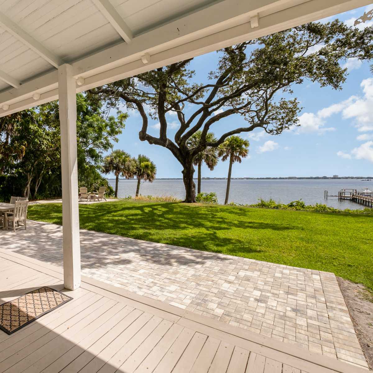 A covered patio with wooden flooring overlooking a grassy area and a river, surrounded by trees and palm plants.