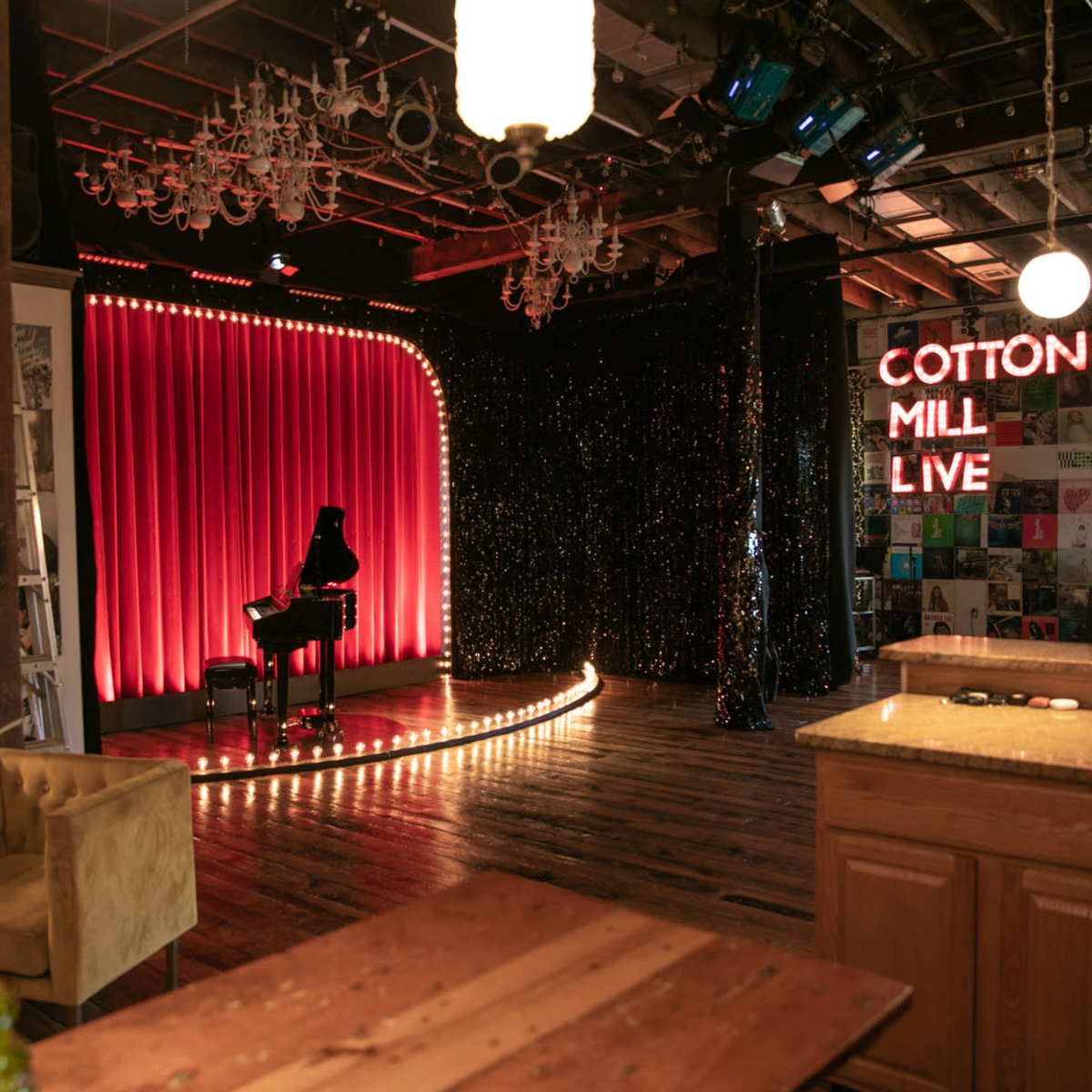 A performance stage at Cotton Mill Live, featuring a red curtain, a piano, and illuminated signage, with a cozy seating area visible in the foreground.