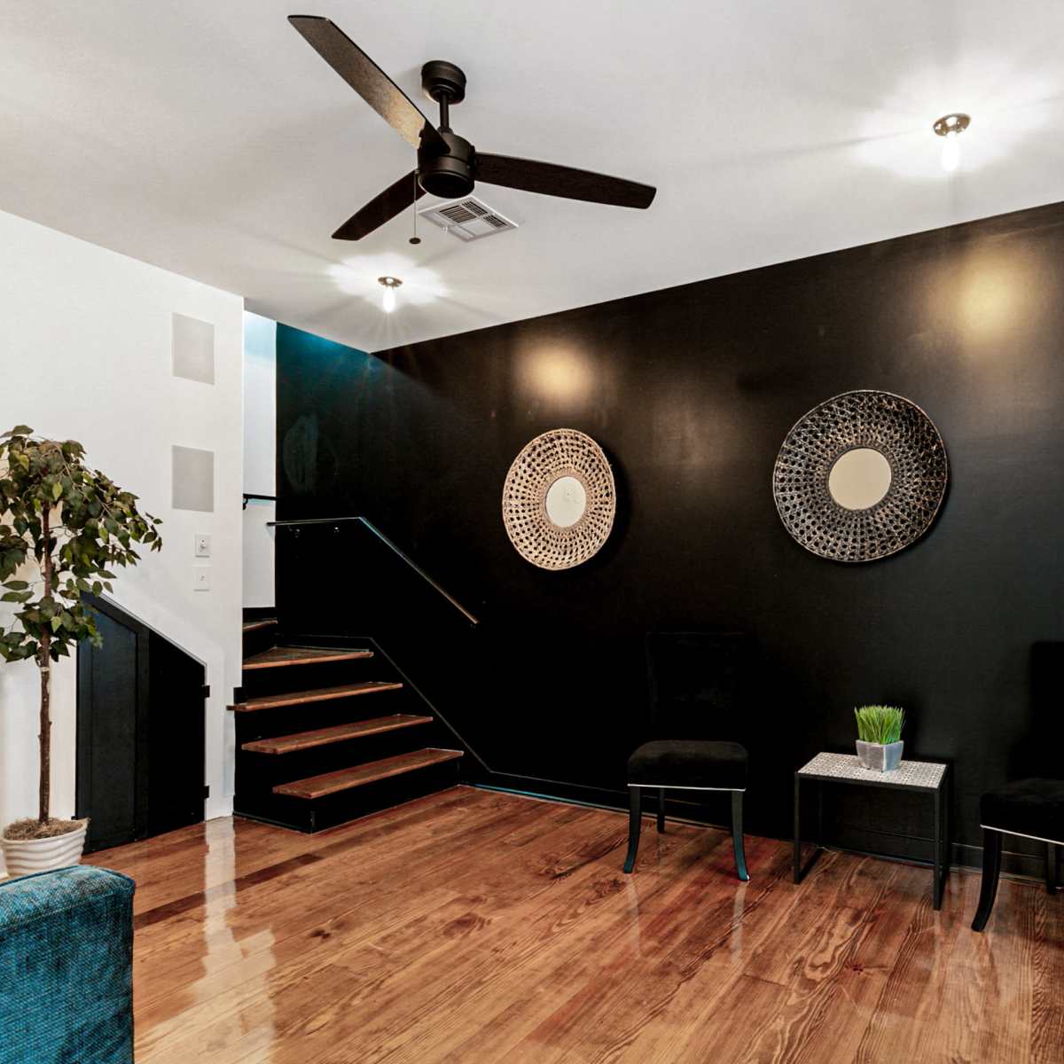 A modern interior space with dark walls, wooden floors, two black chairs, a small table with a plant, and a staircase leading to an upper level.
