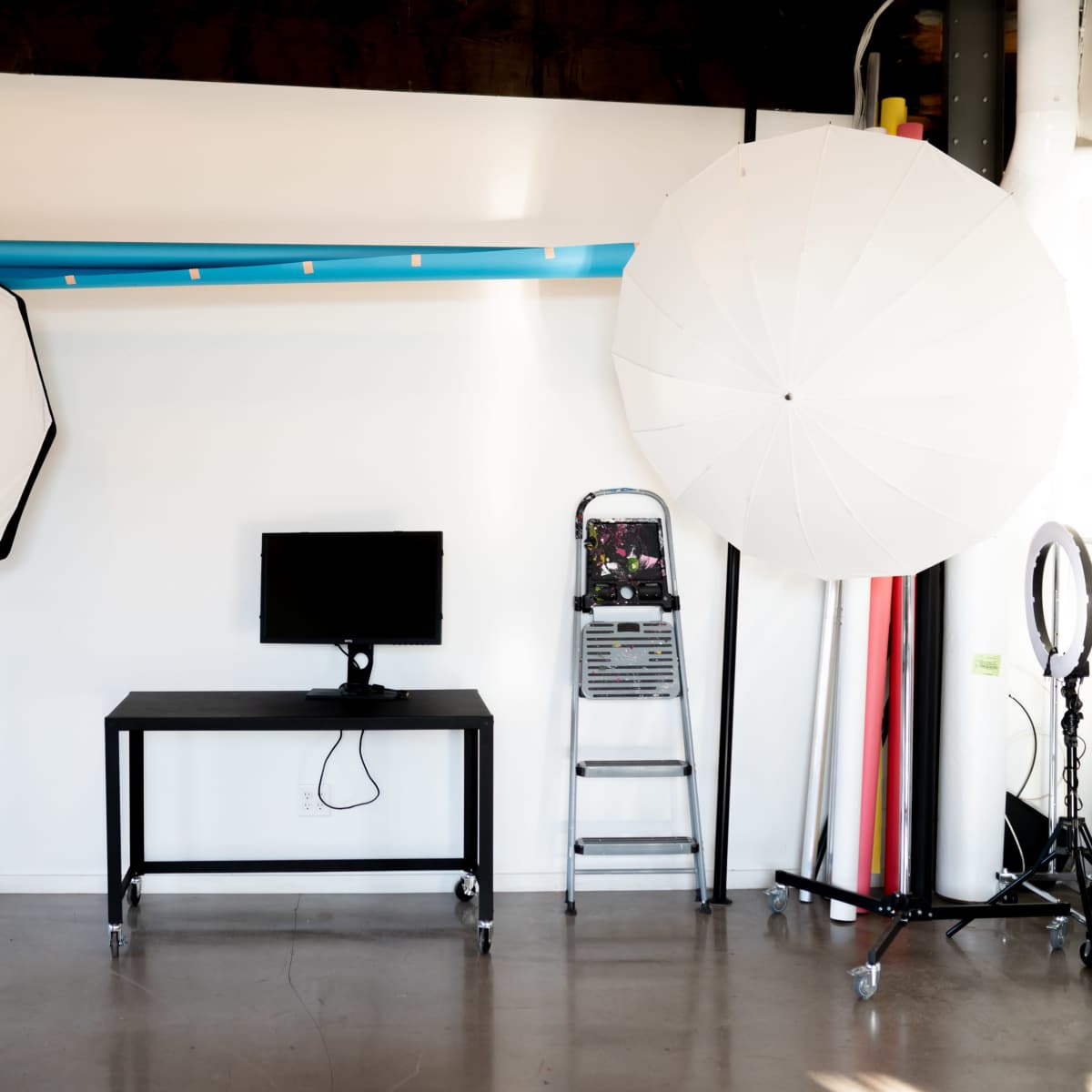 A studio setup featuring two large softbox lights, a black table with a monitor, a ladder, and several rolls of backdrop paper against a plain wall.