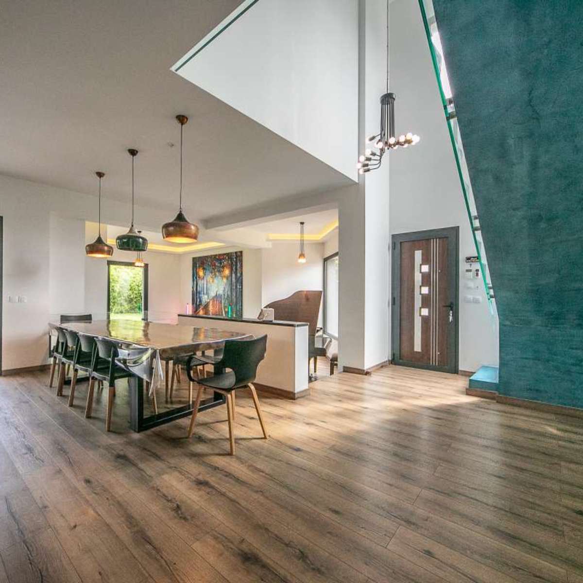 A modern dining area features a large table with black chairs, illuminated by pendant lights, and opens to a garden view through glass doors.