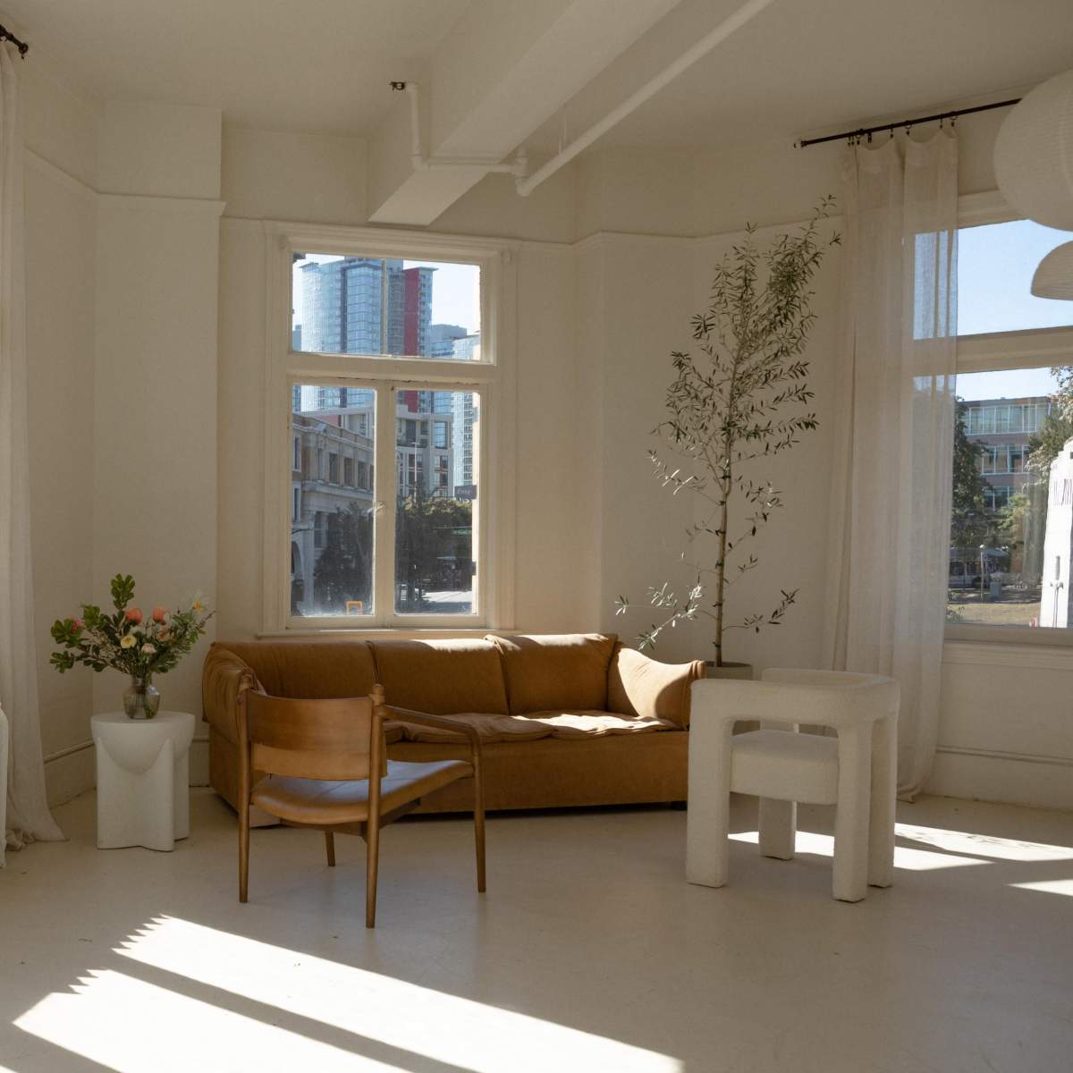 A bright, minimalist interior features a brown couch, a wooden chair, and a white table beside large windows with soft curtains.
