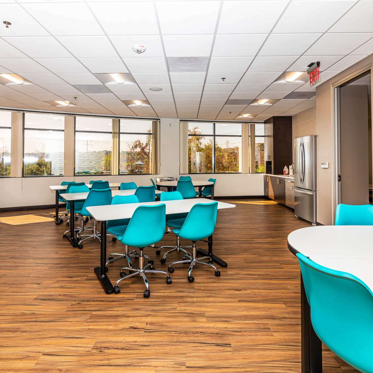 A brightly lit meeting room with several round tables and turquoise chairs, surrounded by large windows.