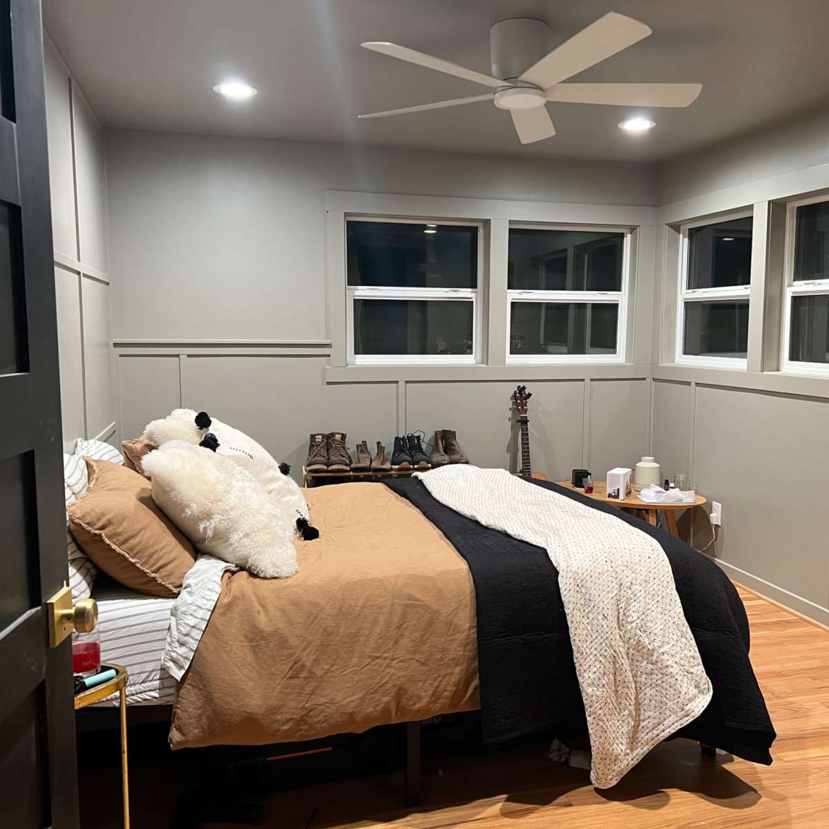 A bedroom featuring a bed with brown and black bedding, wooden flooring, and several windows allowing light to enter.