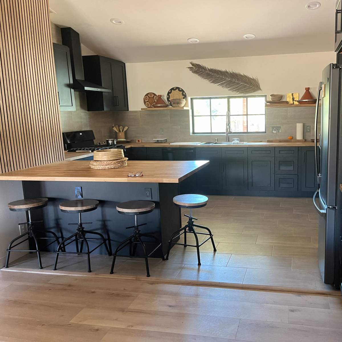 A modern kitchen featuring black cabinets, a wooden countertop with three stools, and a large window allowing natural light.
