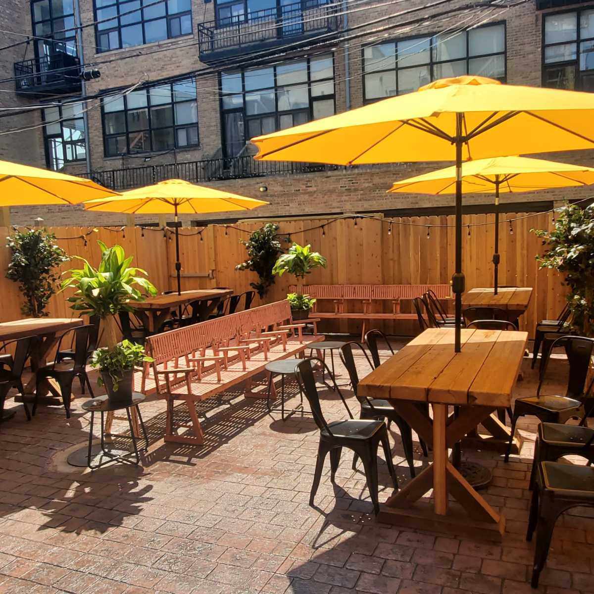 A courtyard with several wooden tables and yellow umbrellas surrounded by wooden fencing and greenery.