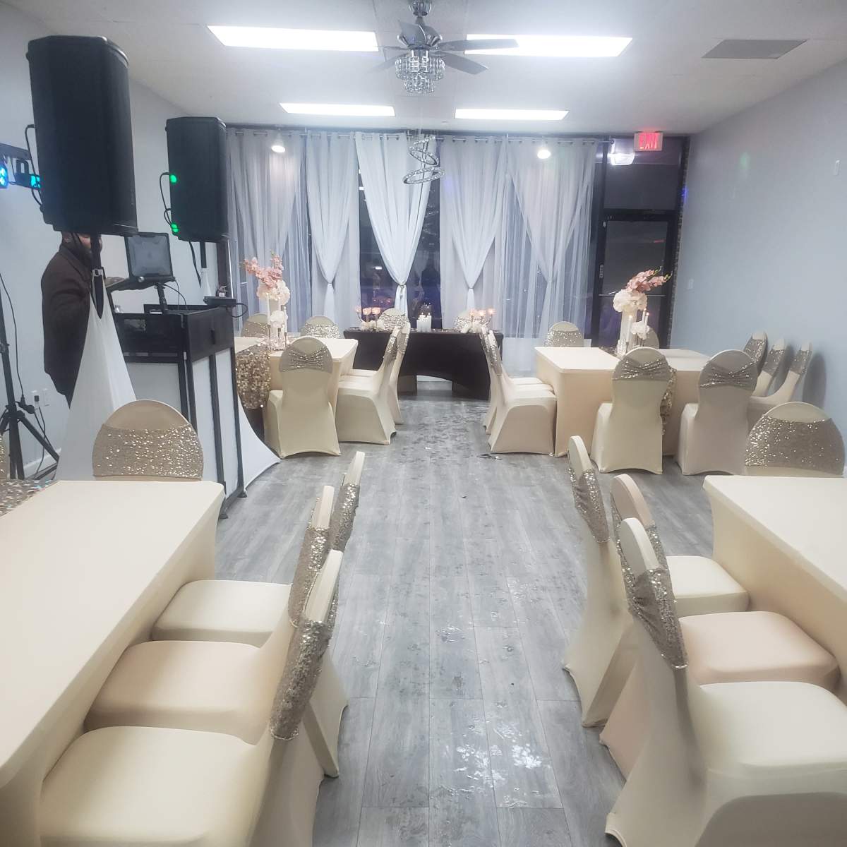 A spacious event room features neatly arranged tables with beige tablecloths and decorated chairs, illuminated by soft lighting and adorned with floral centerpieces.