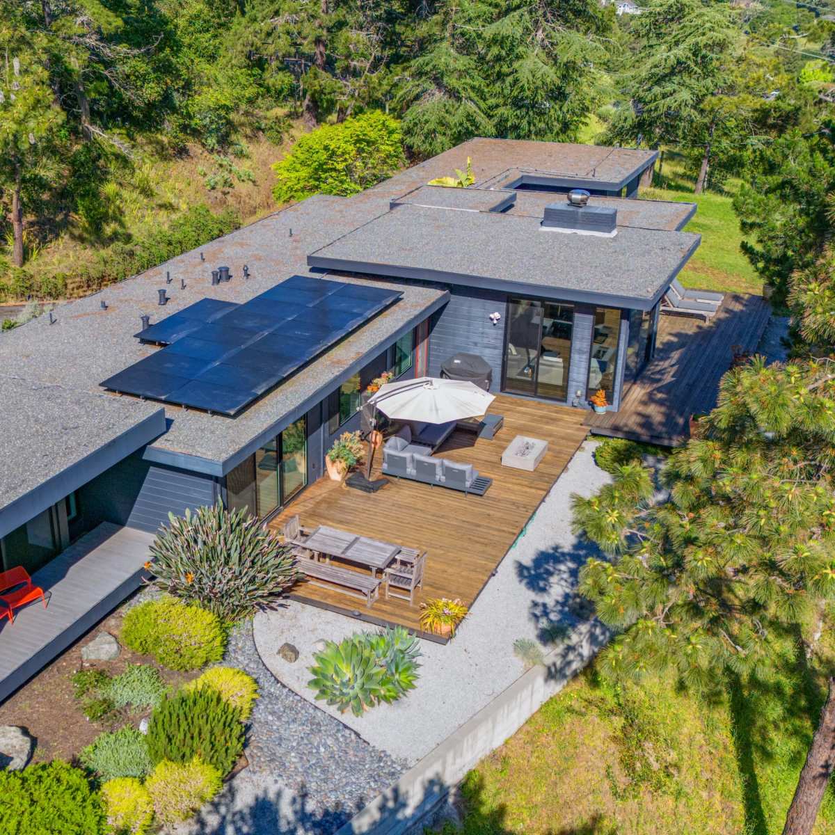 A modern single-story home features a flat roof with solar panels, surrounded by landscaped gardens and a wooden deck with outdoor seating.