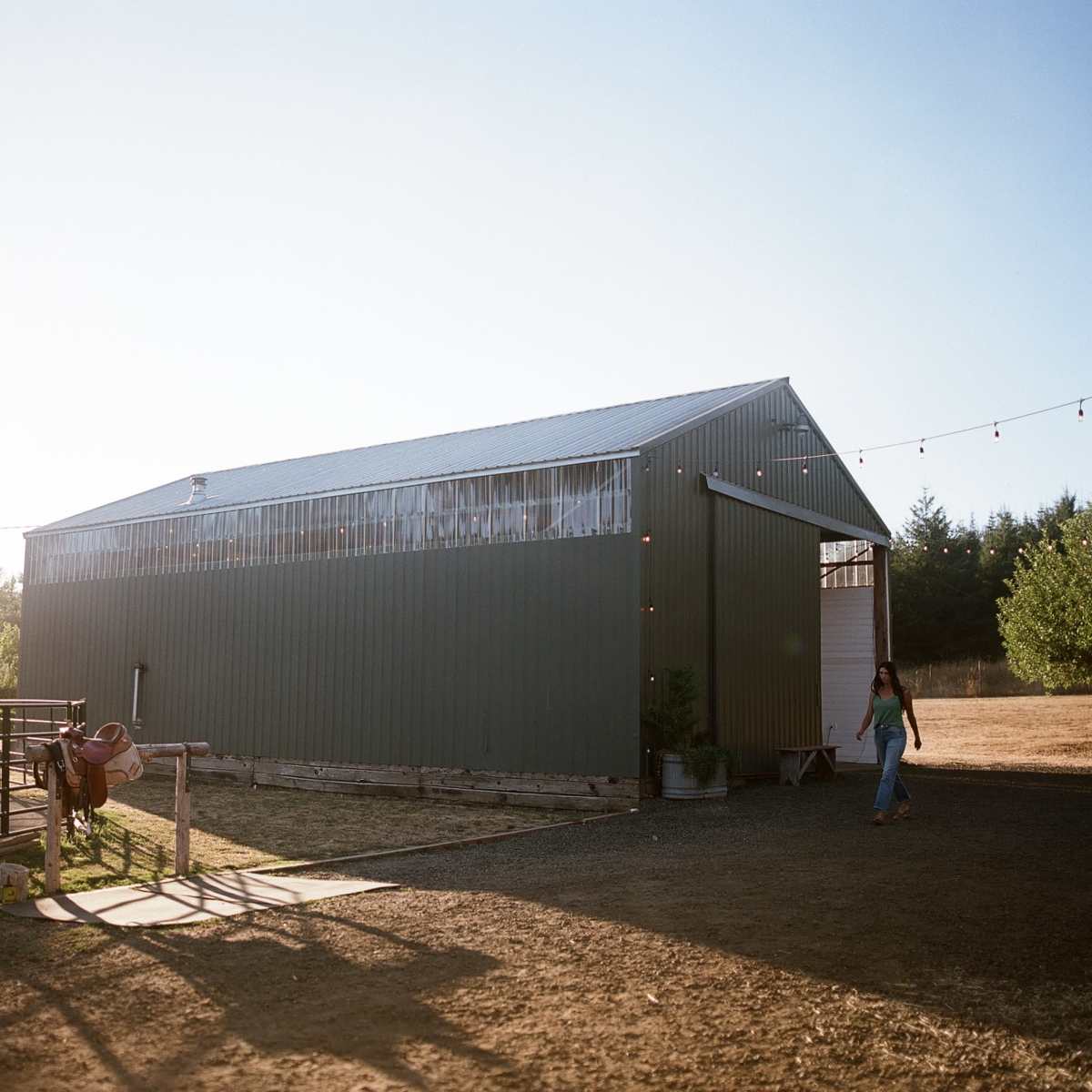A metal barn stands in a rural landscape under a clear blue sky, with a person walking toward it.