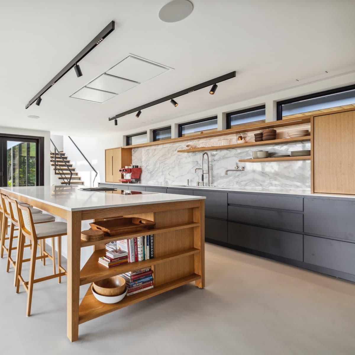 A modern kitchen features a central island with wooden shelving, sleek cabinetry, and large glass doors opening to an outdoor space.