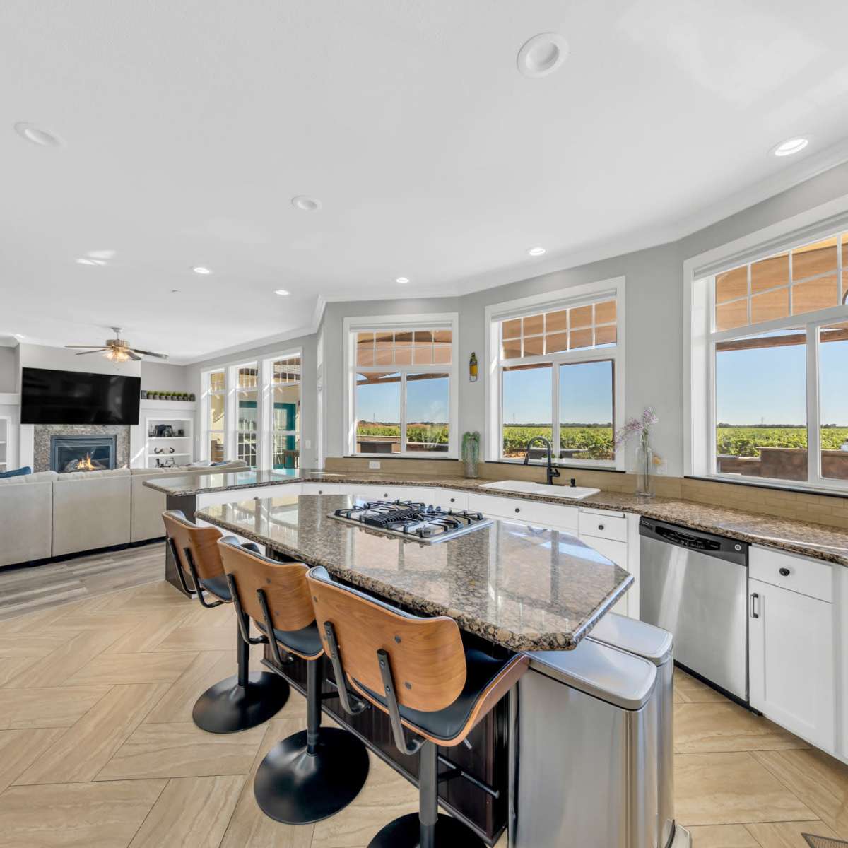A modern kitchen with a large island, equipped with bar stools, overlooking a spacious living area and scenic outdoor views through multiple windows.