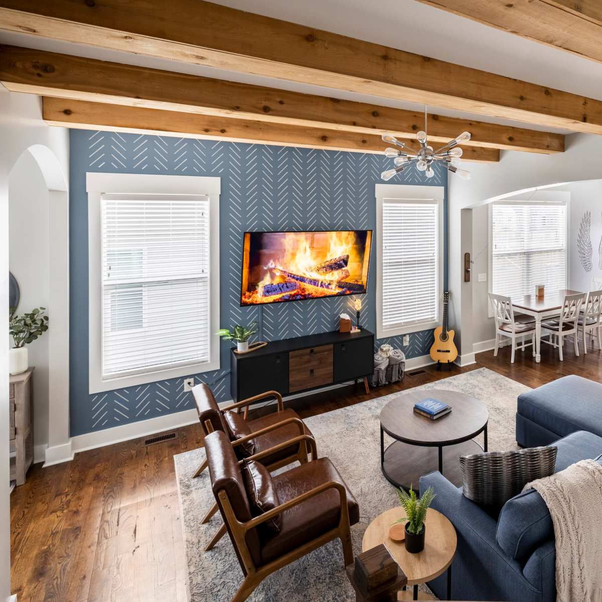 A spacious living room features a blue sectional couch, wooden beams on the ceiling, a television mounted above a dark cabinet, and a cozy fireplace.