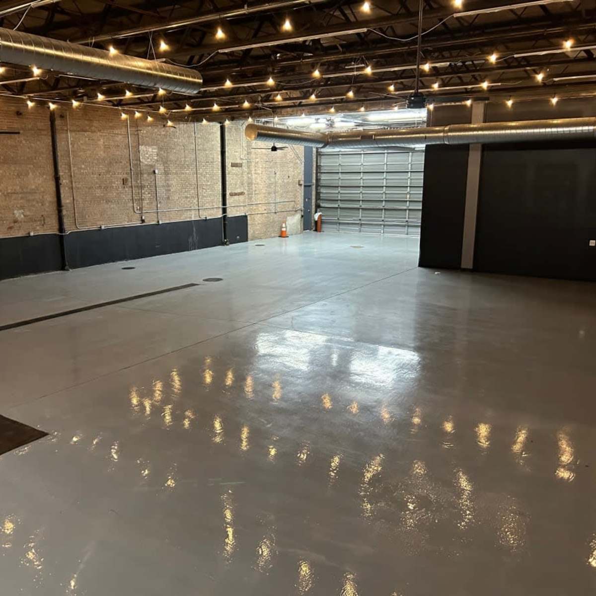 A large, empty industrial space with a polished gray concrete floor and exposed brick walls, featuring overhead lighting and a garage door.