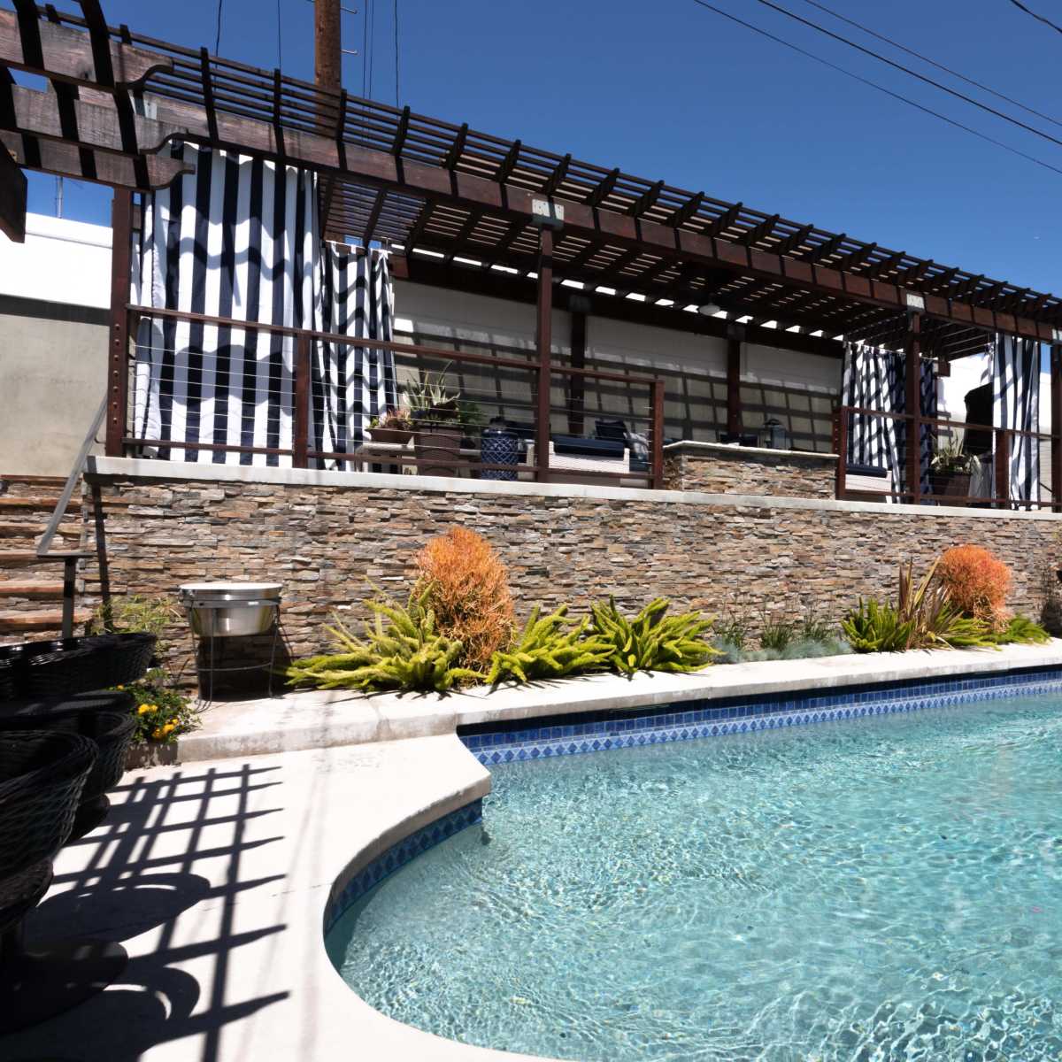 A clear blue swimming pool is surrounded by stonework and landscaped plants, with a wooden pergola and a patio area in the background.