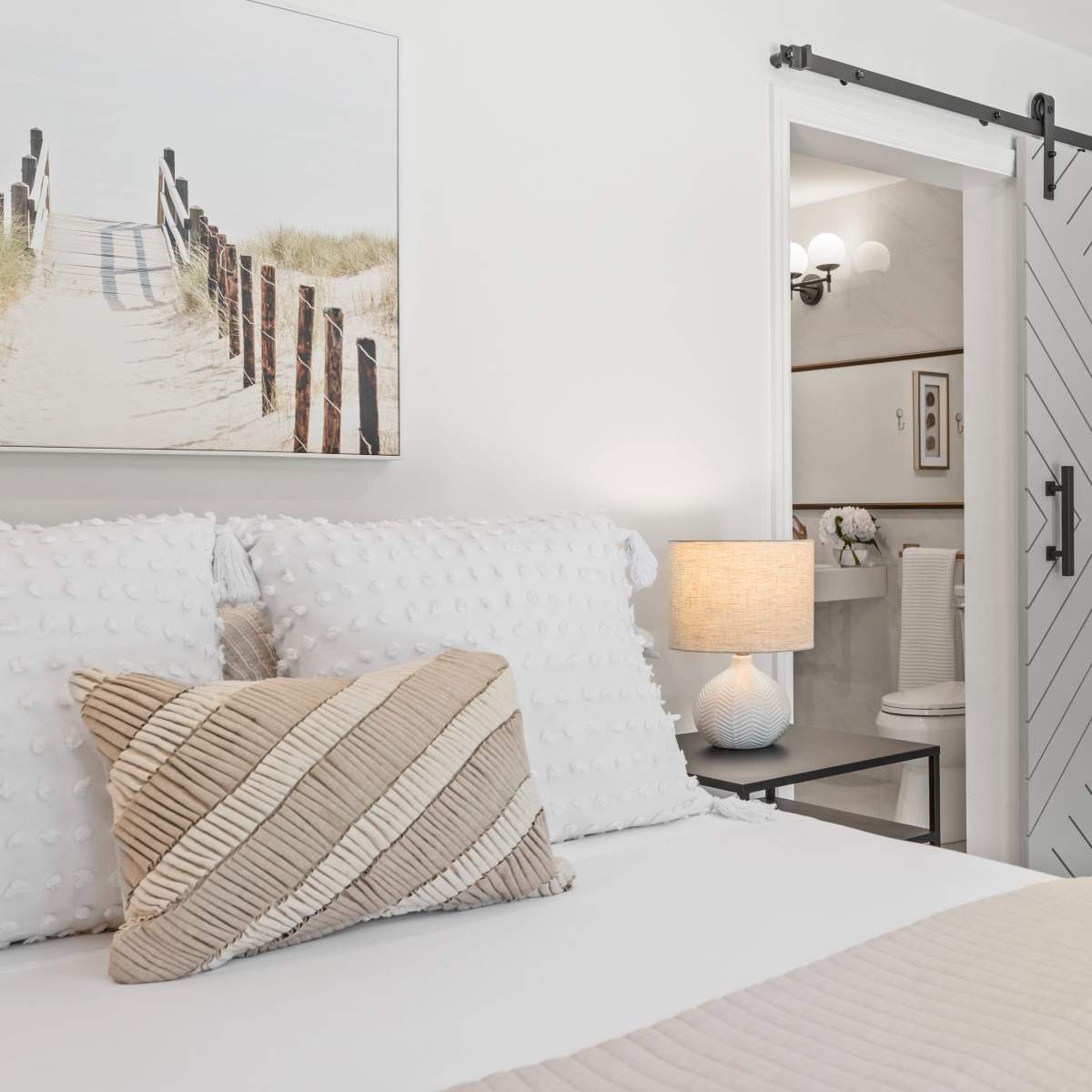 A neatly arranged bedroom features a bed with decorative pillows, a bedside lamp, and a sliding barn door leading to a bathroom.