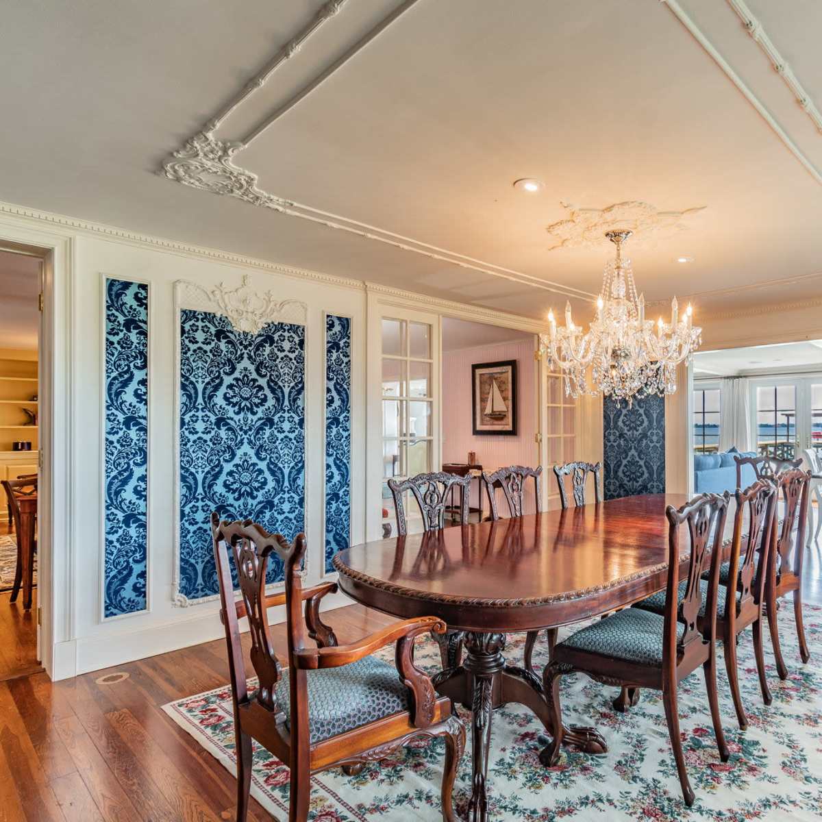 The dining room features a large wooden table surrounded by twelve chairs, illuminated by a chandelier and accented by blue patterned wallpaper and floral carpeting.
