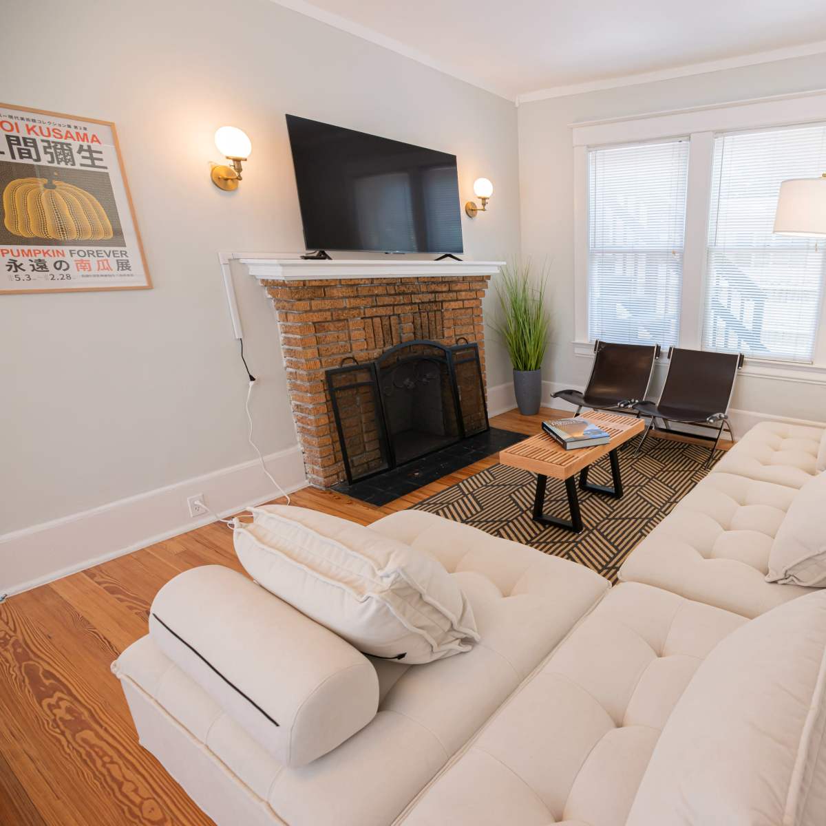 A living room featuring a sectional sofa, a TV mounted above a brick fireplace, and two lounge chairs beside a coffee table.