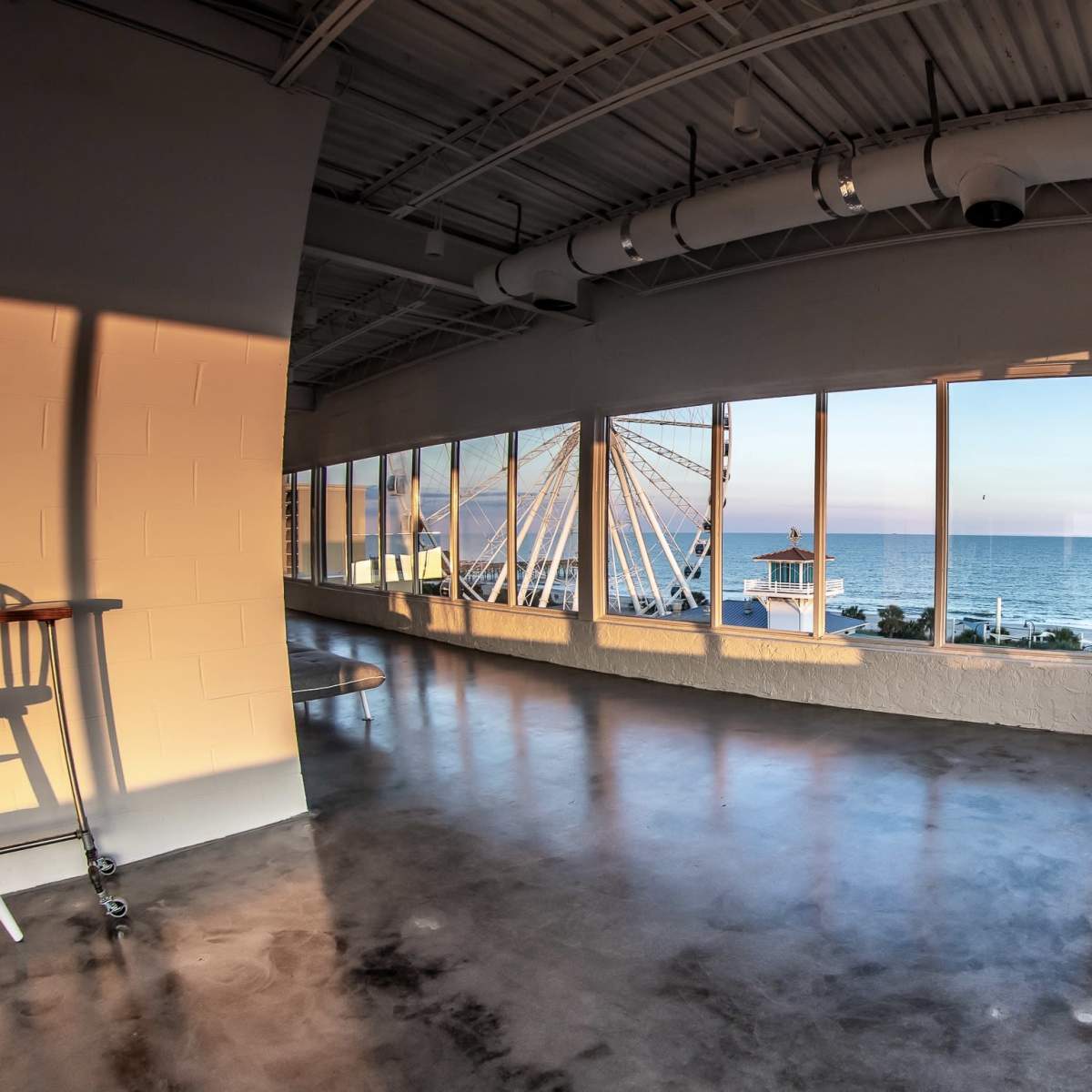 A modern, empty room with large windows overlooking a beach and a Ferris wheel in the distance.