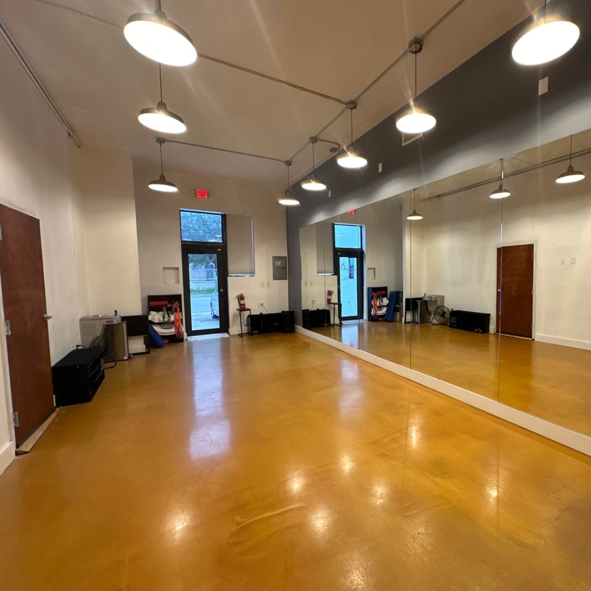 A spacious, well-lit room with a large mirror on one wall, hardwood flooring, and exit doors.