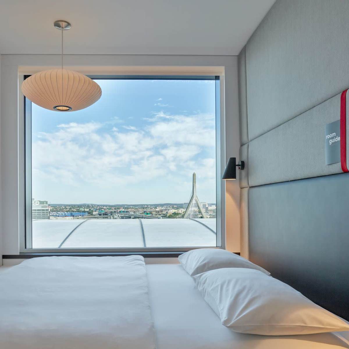 A modern bedroom features a large window with a city view, a wall-mounted television, and a minimalist design with a bed and a pendant light.