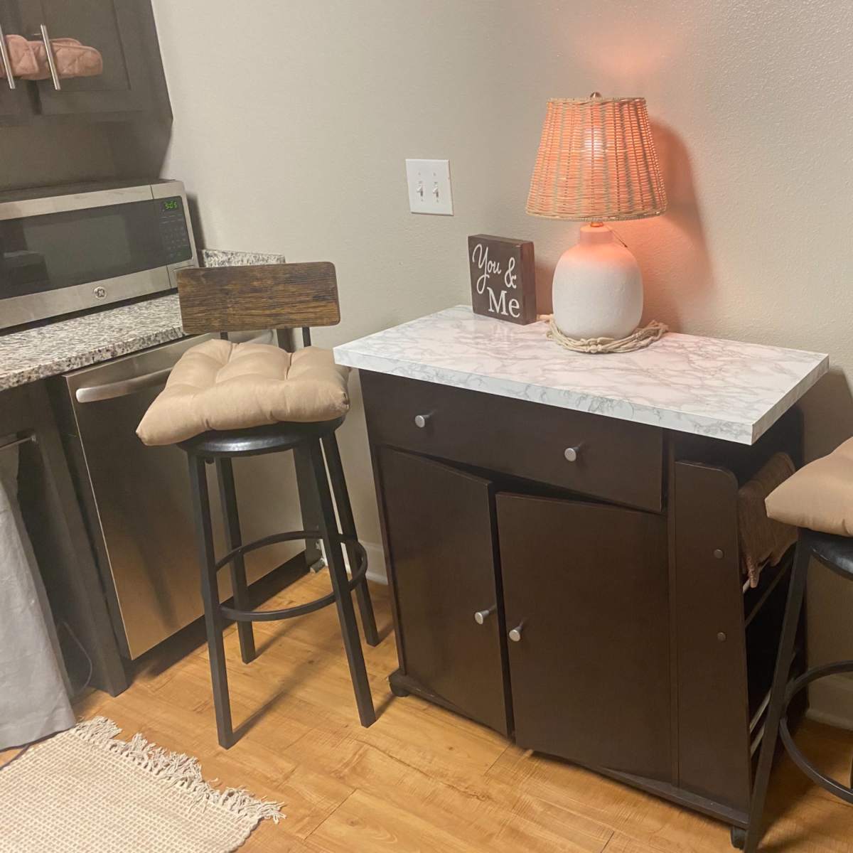 A small kitchen area featuring a side table with a marble top, two black stools with cushions, and a lamp with a woven shade.