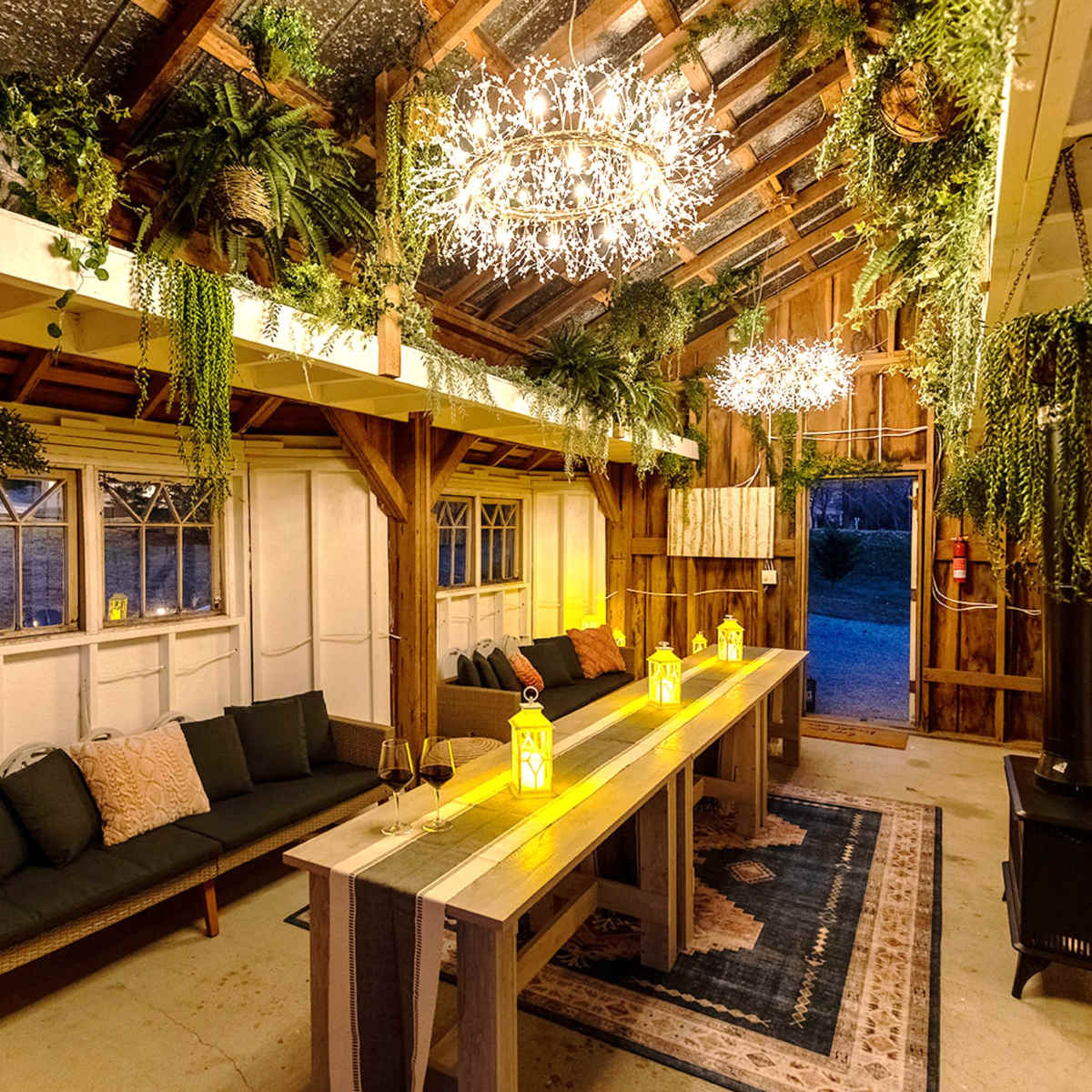 A rustic indoor space featuring a long wooden table, two stylish seating areas, and hanging lights amidst an array of plants.