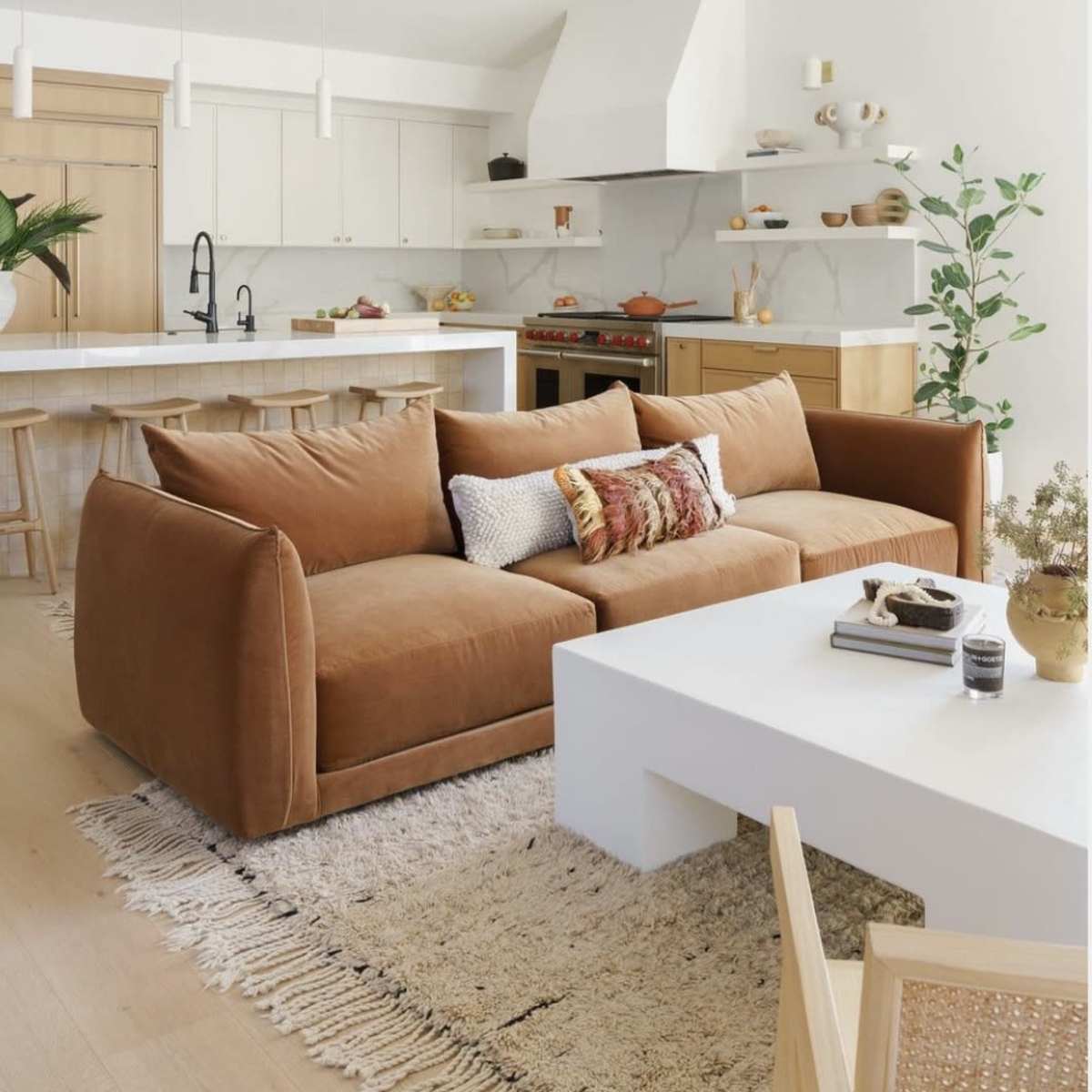 A modern living room featuring a brown sofa, a white coffee table, a light-colored rug, and a kitchen in the background with white cabinets and wood accents.
