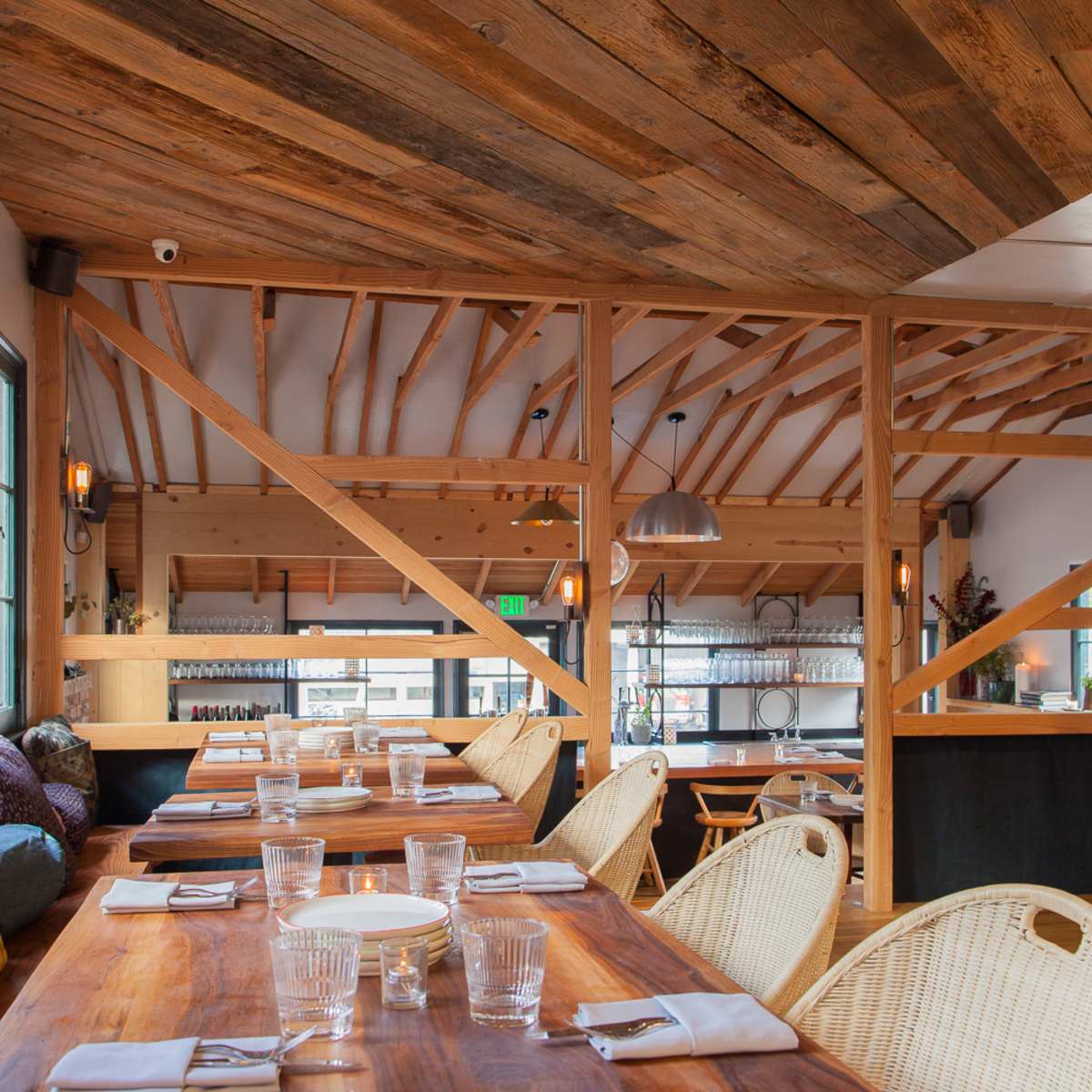 A spacious restaurant interior features wooden beams, dining tables set with plates and glasses, and a bright atmosphere enhanced by large windows.