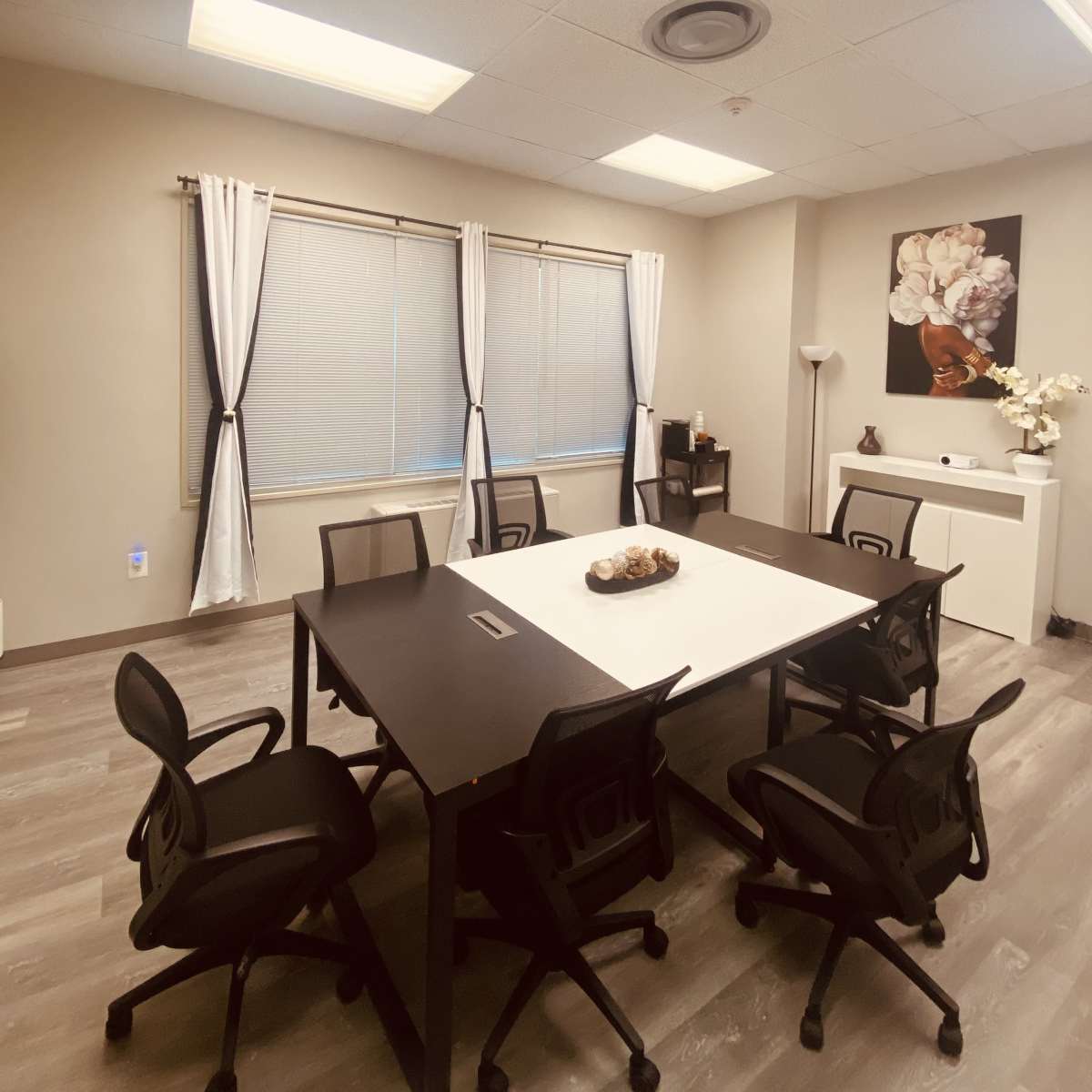 A modern meeting room features a large table surrounded by four chairs, with windows dressed in white curtains and a minimalist decor.