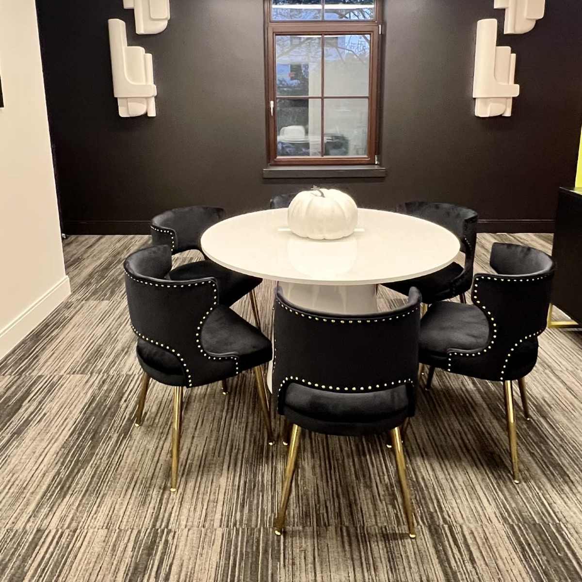 A round table with a white top is surrounded by six black chairs with gold legs in a room featuring dark walls and two decorative light fixtures.