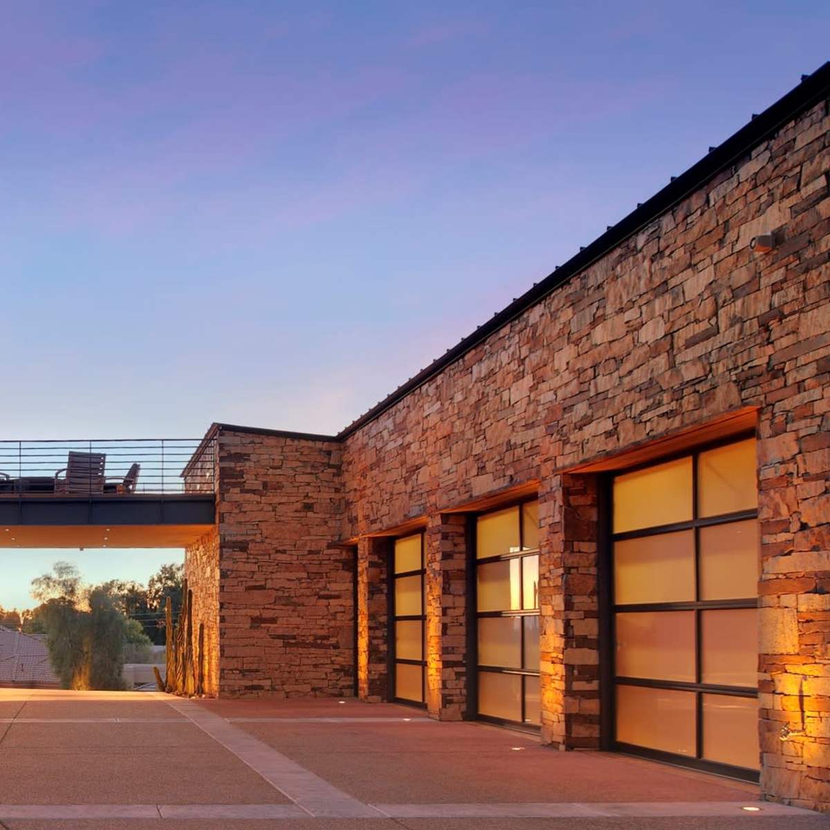 A modern stone and glass house features a bridge connecting two sections, with a driveway leading up to it at dusk.