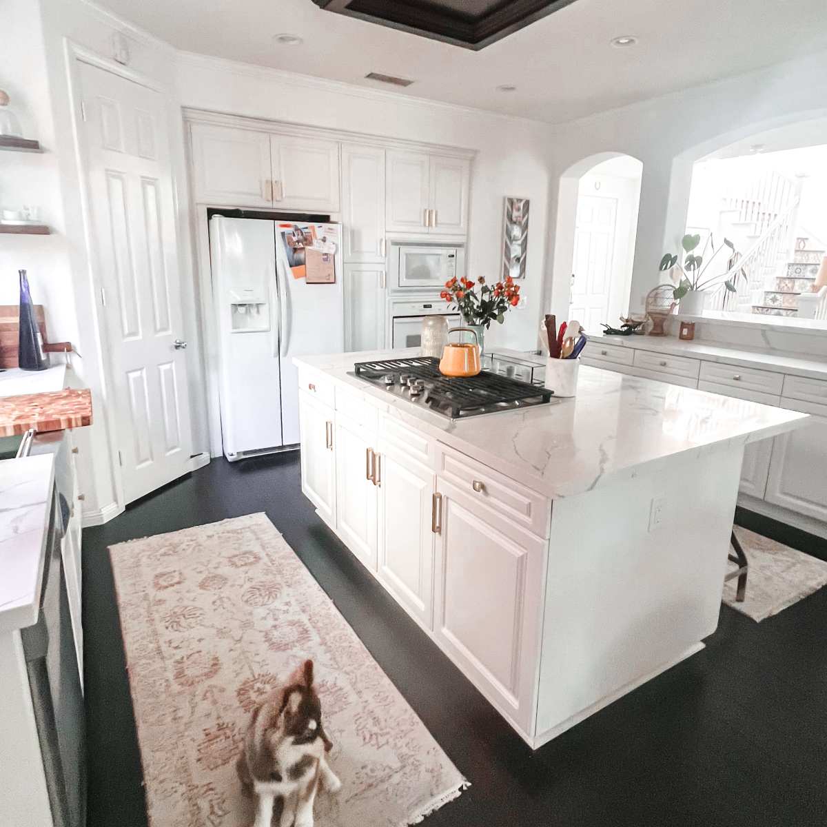 A modern kitchen features a large island with a gas stove, white cabinetry, and a pet dog on a floral rug.