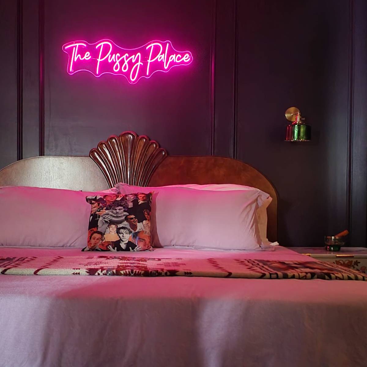 A bed with a decorative headboard is centered against a dark wall, featuring a neon sign reading "The Pussy Palace" above it.