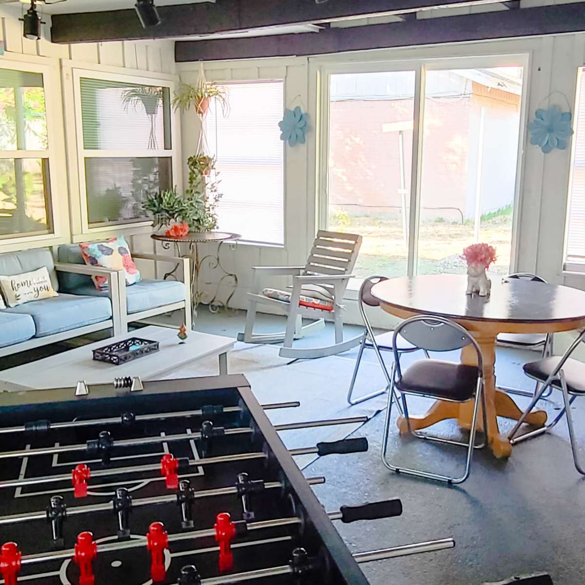 A screened-in porch with a seating area, a foosball table, and a dining table surrounded by chairs.