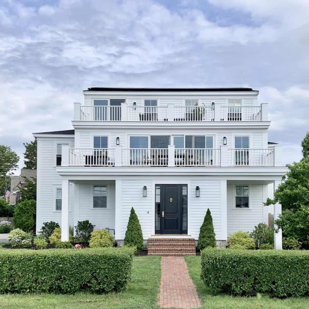 A three-story white house with balconies on each floor is flanked by neatly trimmed hedges and a brick pathway leading to the front door.