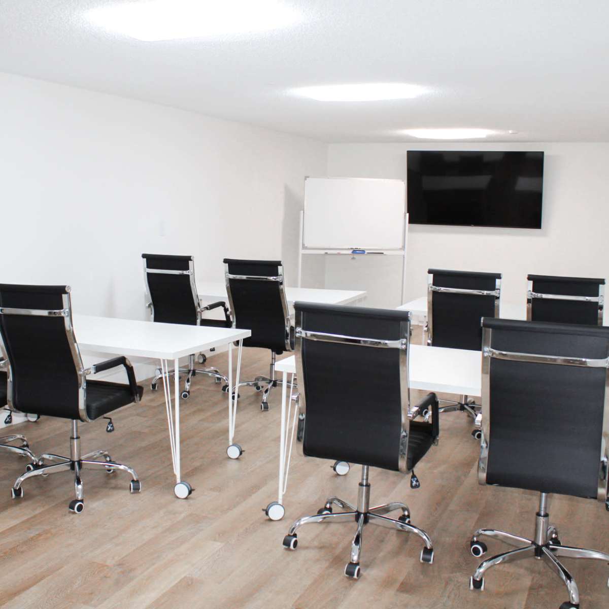 A minimalist conference room features several black rolling chairs around white tables, with a whiteboard and a television mounted on the wall.