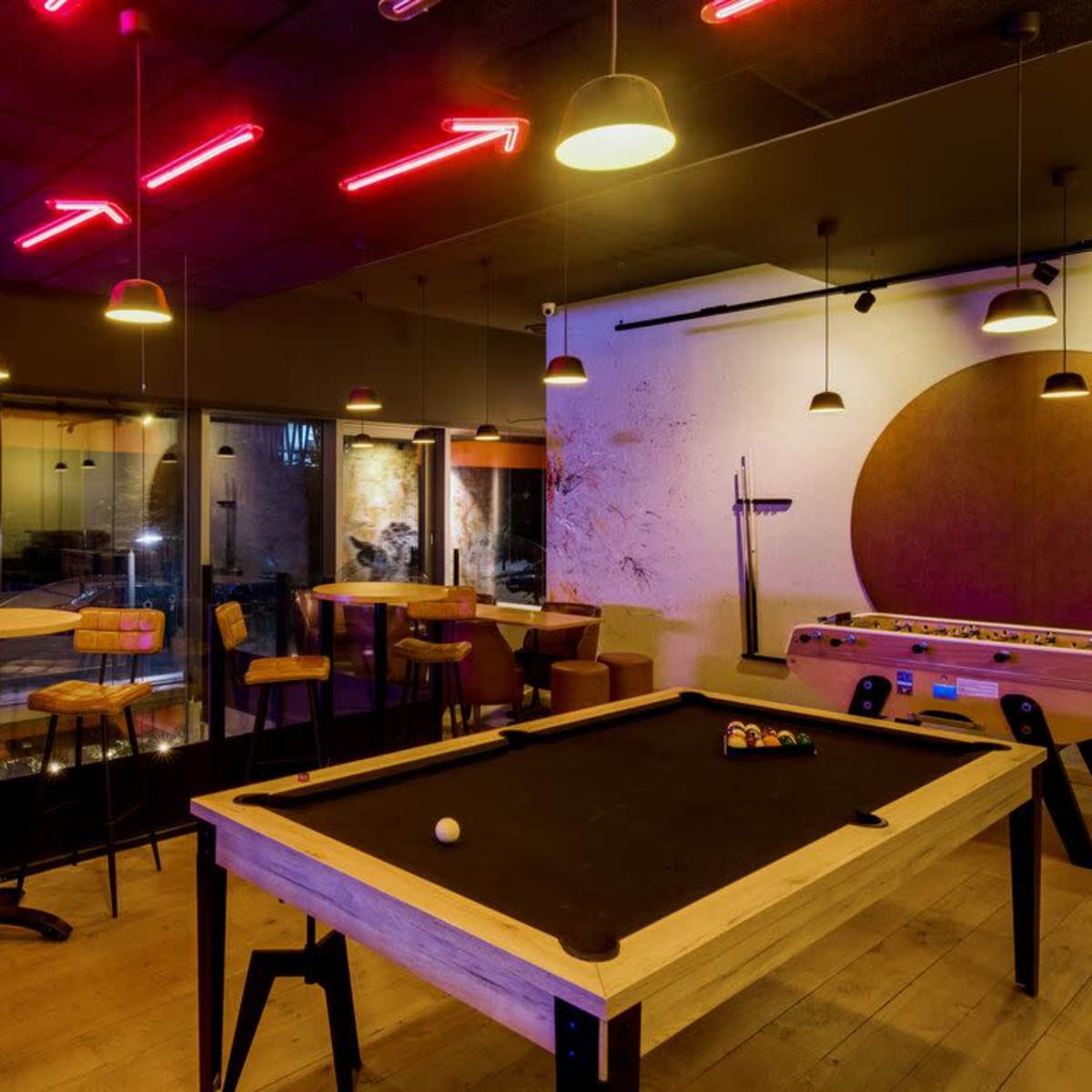 A game room with a pool table, foosball table, and neon lighting, featuring wooden stools and tables against a backdrop of large windows.
