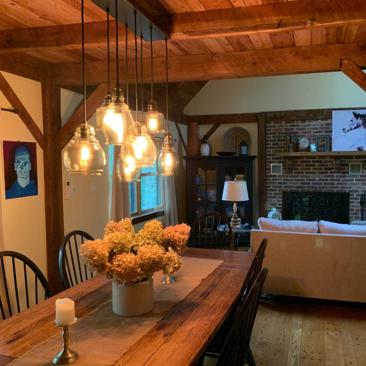 A rustic dining area with a wooden table, hanging light fixtures, and a view of a cozy living room featuring a brick fireplace and a beige sofa.