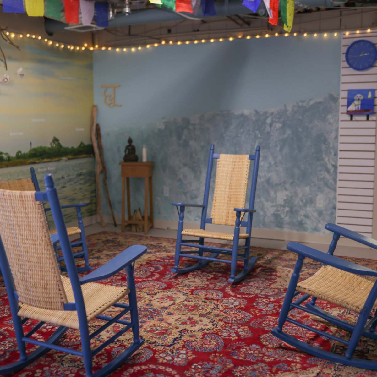 A cozy room features four blue rocking chairs arranged on a patterned rug, with a wall mural depicting a paddleboarder and strings of lights above.