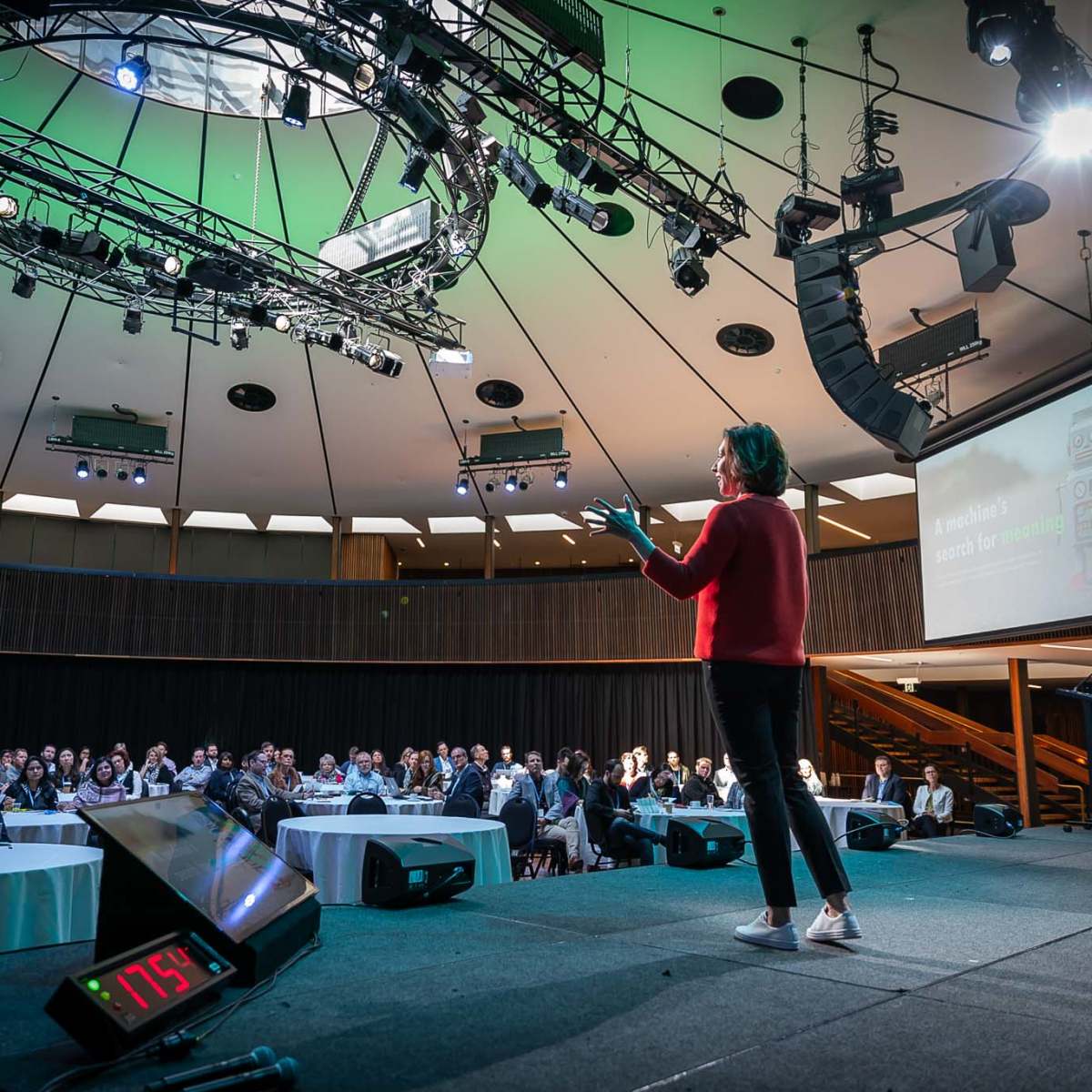 A speaker presents on stage in a well-lit auditorium filled with an audience seated at tables.