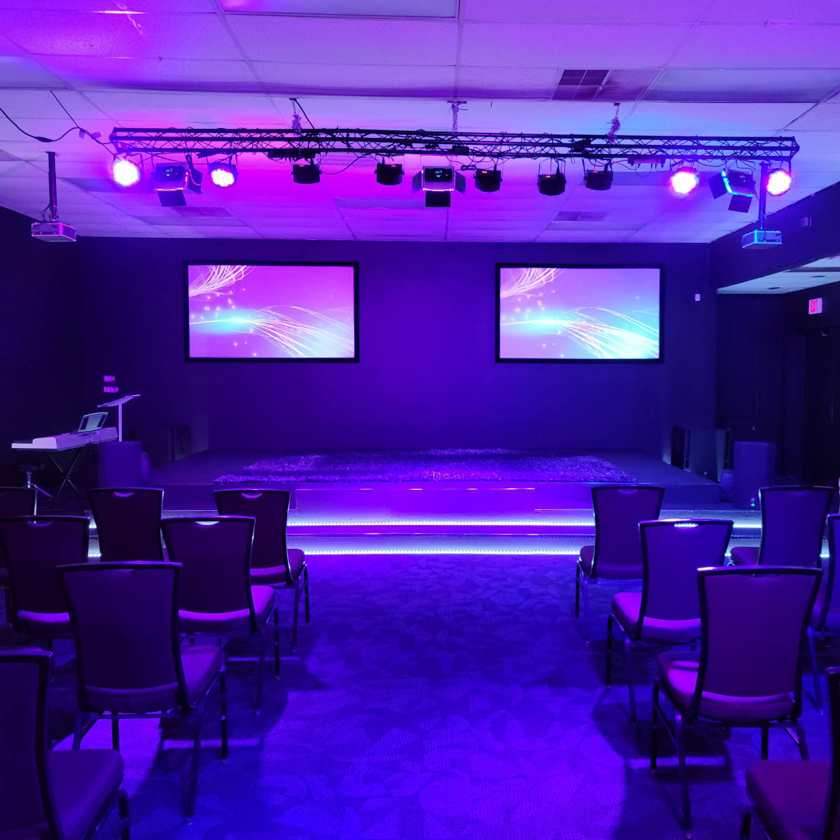 A dimly lit event space with purple lighting, featuring two screens on the wall, a small stage, and rows of chairs facing the stage.