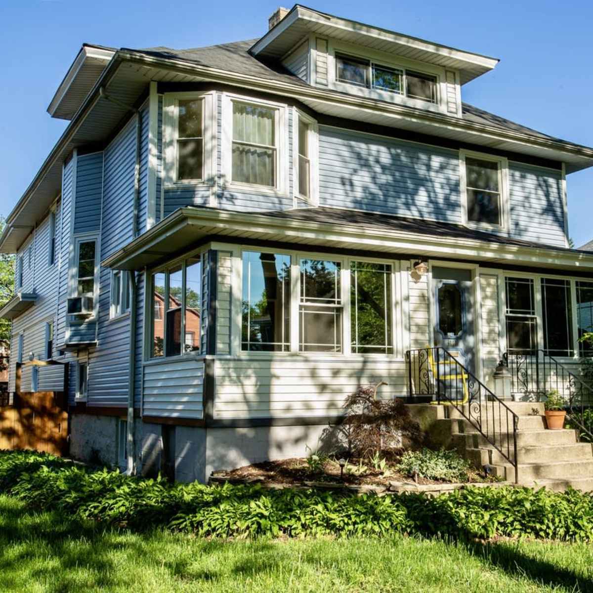 A three-story house with a porch and large windows is surrounded by greenery and a well-kept lawn.