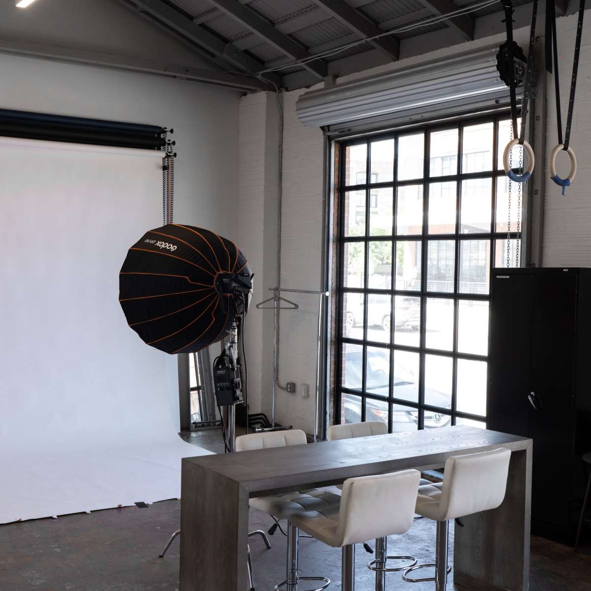 A photography studio with a white backdrop, a large light source, and a table with four chairs near a large window.